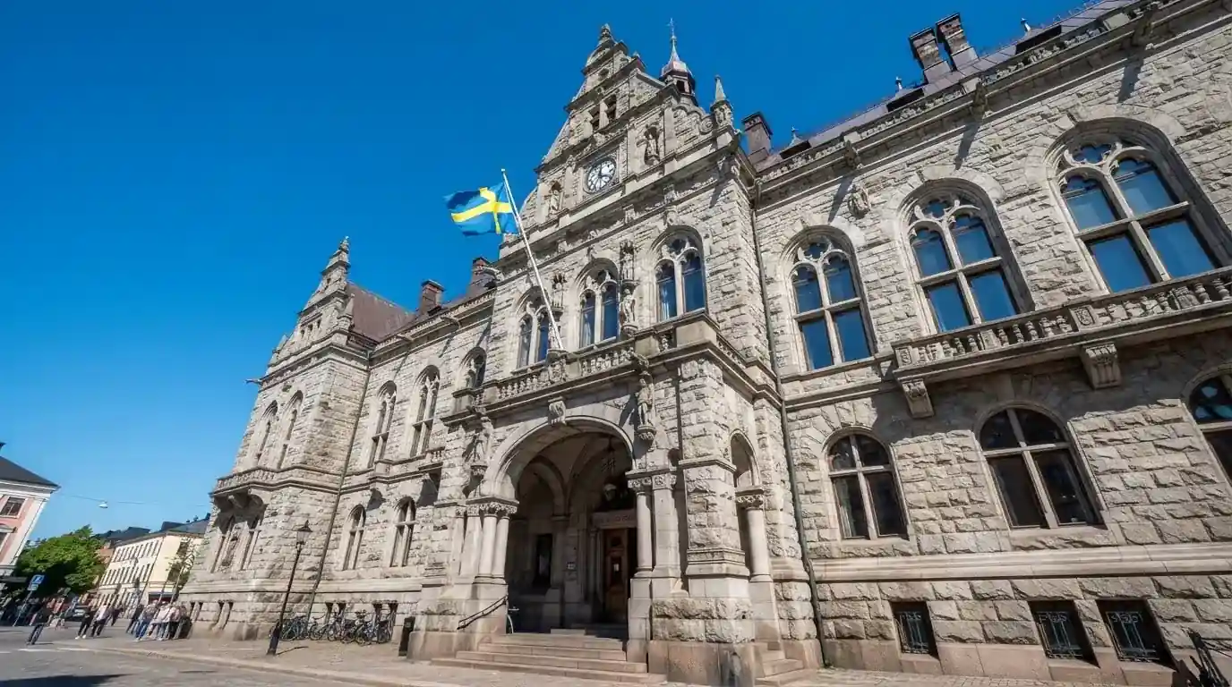 Ett gammalt stadshus i sten fotograferat underifrån mot en klarblå himmel, med den svenska flaggan som fladdrar ovanför entrén.
